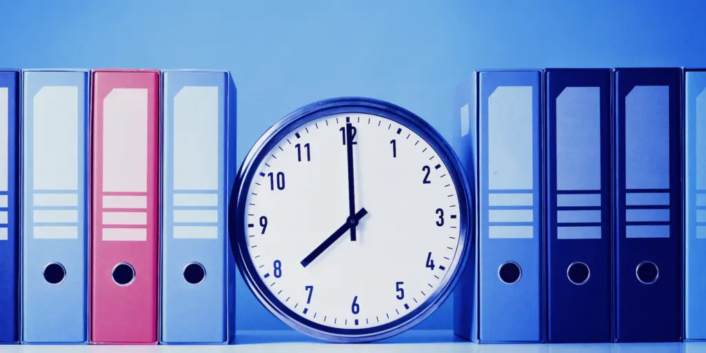 A wall clock sitting next to a row of binders. The clock is a black and silver round analog clock with Roman numerals.